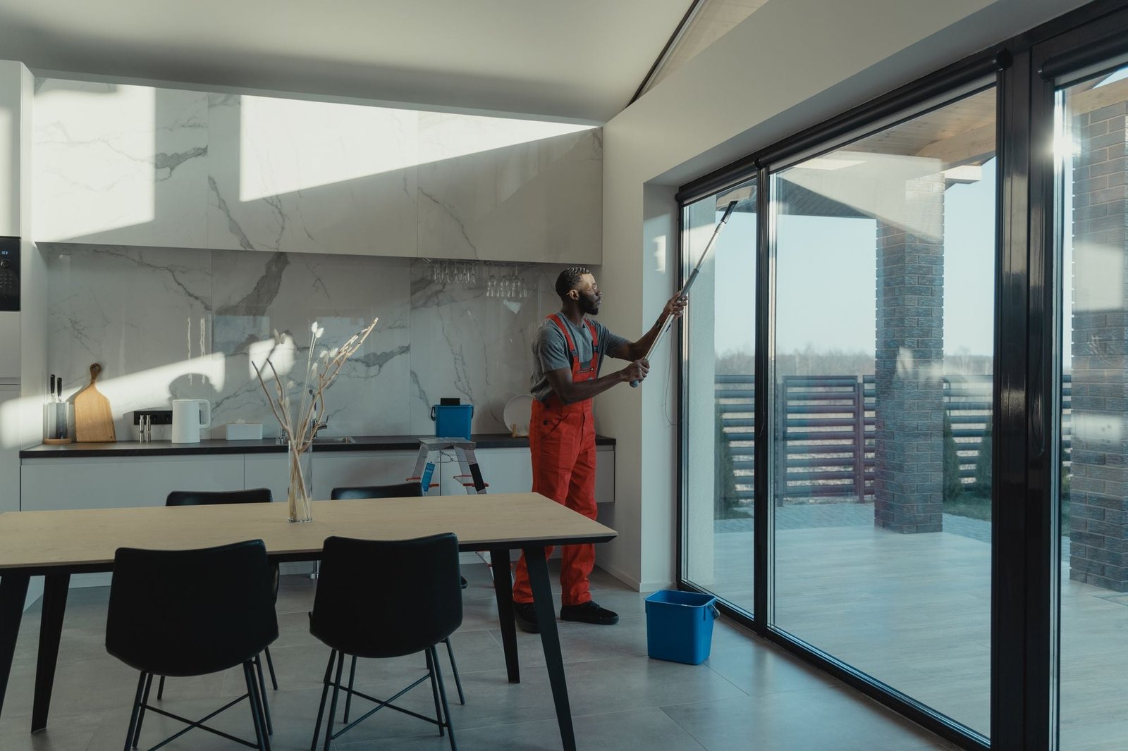 Een schoonmaker reinigt met een lange steel een grote glazen schuifpui naast een keuken