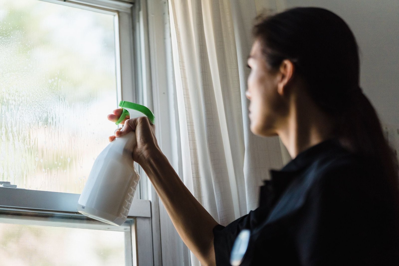 Een vrouw spuit glasreiniger op een raam naast een gordijn